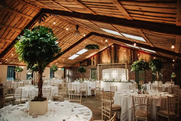 Rustic barn interior with high wooden beams, elegantly set round tables, and lush greenery, creating a charming wedding atmosphere.
