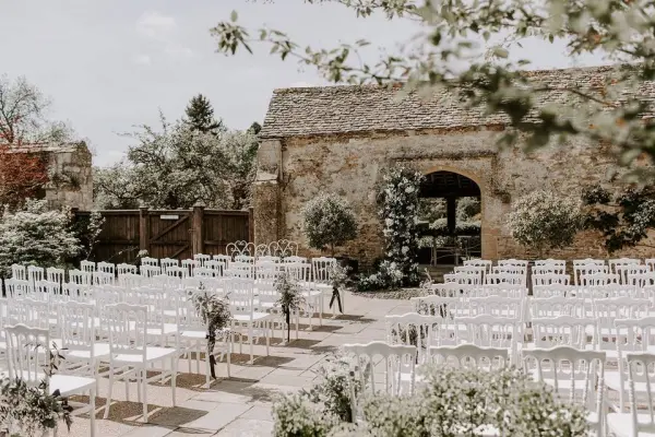 Charming outdoor ceremony space featuring white chairs, lush greenery, and a rustic stone backdrop for a romantic wedding setting.