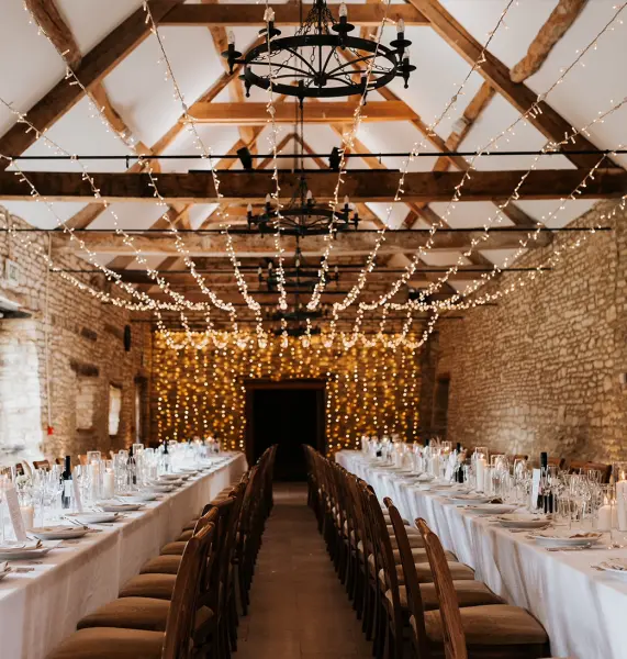 Rustic banquet hall adorned with string lights and wooden beams, featuring long tables set for an elegant wedding reception.