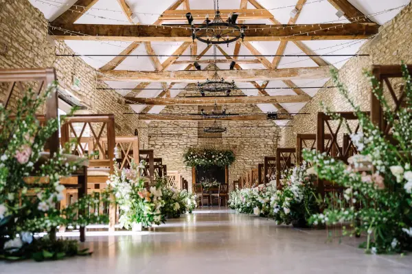 Charming stone-walled ceremony space featuring wooden beams, elegant seating, and abundant floral arrangements lining the aisle.
