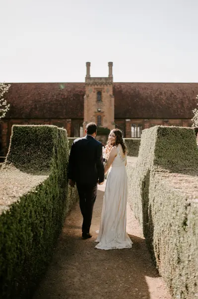 Couple walking hand-in-hand through lush hedges, leading to a charming historical building, perfect for outdoor wedding ceremonies.