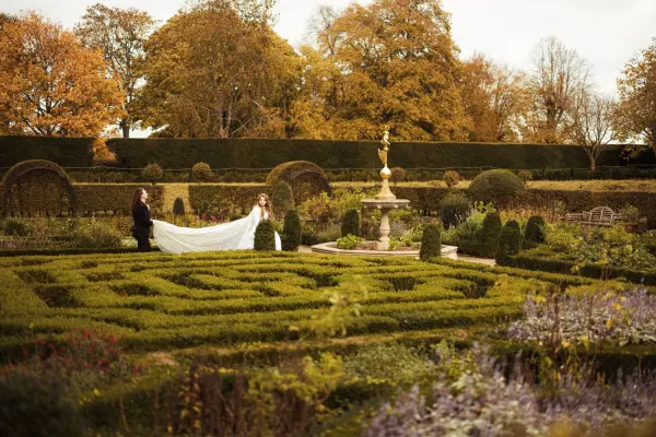Couple in a romantic garden setting, featuring intricate hedges, autumn foliage, and a beautiful fountain, perfect for outdoor ceremonies.