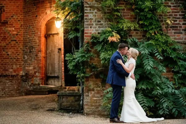 A couple shares a romantic kiss in front of a rustic brick wall adorned with greenery, creating an intimate outdoor setting.