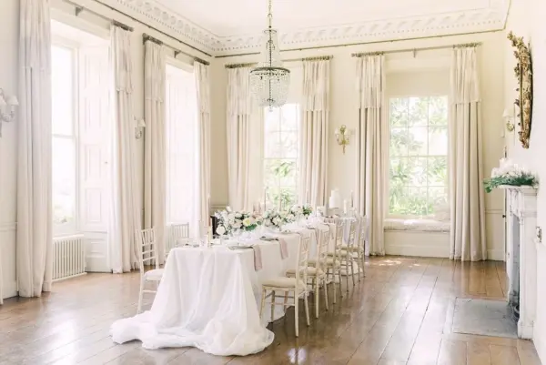 Bright, elegant dining area with large windows, wooden floors, and a refined table set for a wedding reception surrounded by soft drapes.