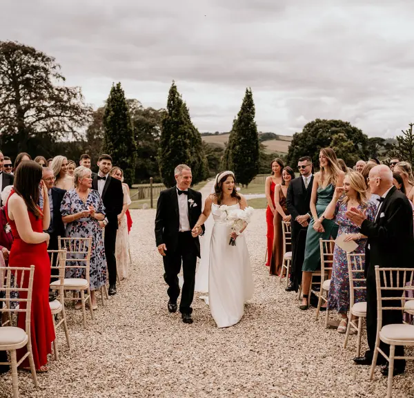 A bride in a flowing white gown walks down a gravel aisle flanked by guests seated on elegant chairs, lush greenery in the background.