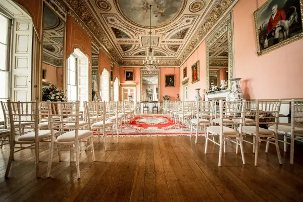 Elegant ceremony space featuring high ceilings, ornate decorations, and polished wooden floors, set with white chiavari chairs.