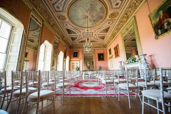 Elegant ceremony room featuring ornate ceilings, classic chandeliers, and rows of chiavari chairs against a backdrop of pastel pink walls.