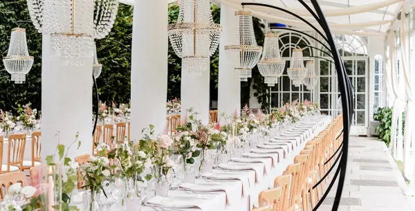 A picturesque dining area features long tables adorned with floral centerpieces, elegant chandeliers, and a lush greenery backdrop.
