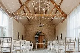 Charming ceremony space featuring exposed wooden beams, soft lighting, and elegant white chairs arranged around a floral-adorned altar.