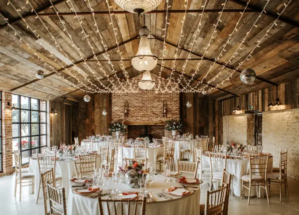 Rustic barn venue featuring wooden beams, elegant chandeliers, and round tables adorned with flowers, perfect for a cozy wedding celebration.