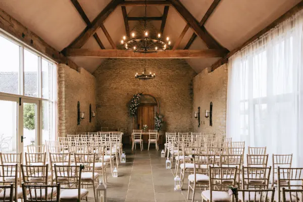 Rustic wedding ceremony space featuring wooden beams, elegant chandeliers, and rows of gold chiavari chairs against a stone wall.
