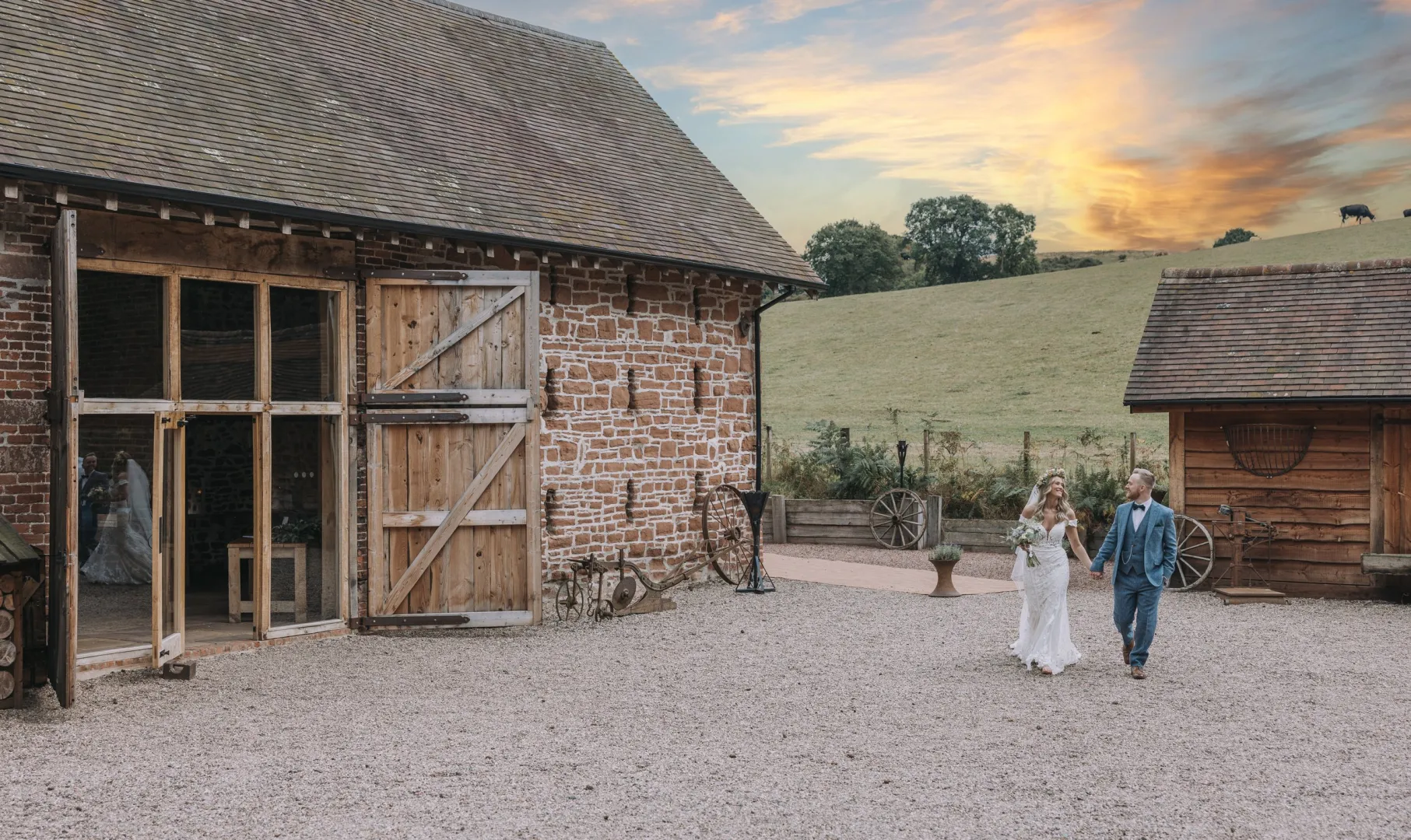 Charming rustic barn backdrop at sunset, with a couple walking hand-in-hand on gravel, surrounded by lush green fields. Perfect for outdoor weddings.