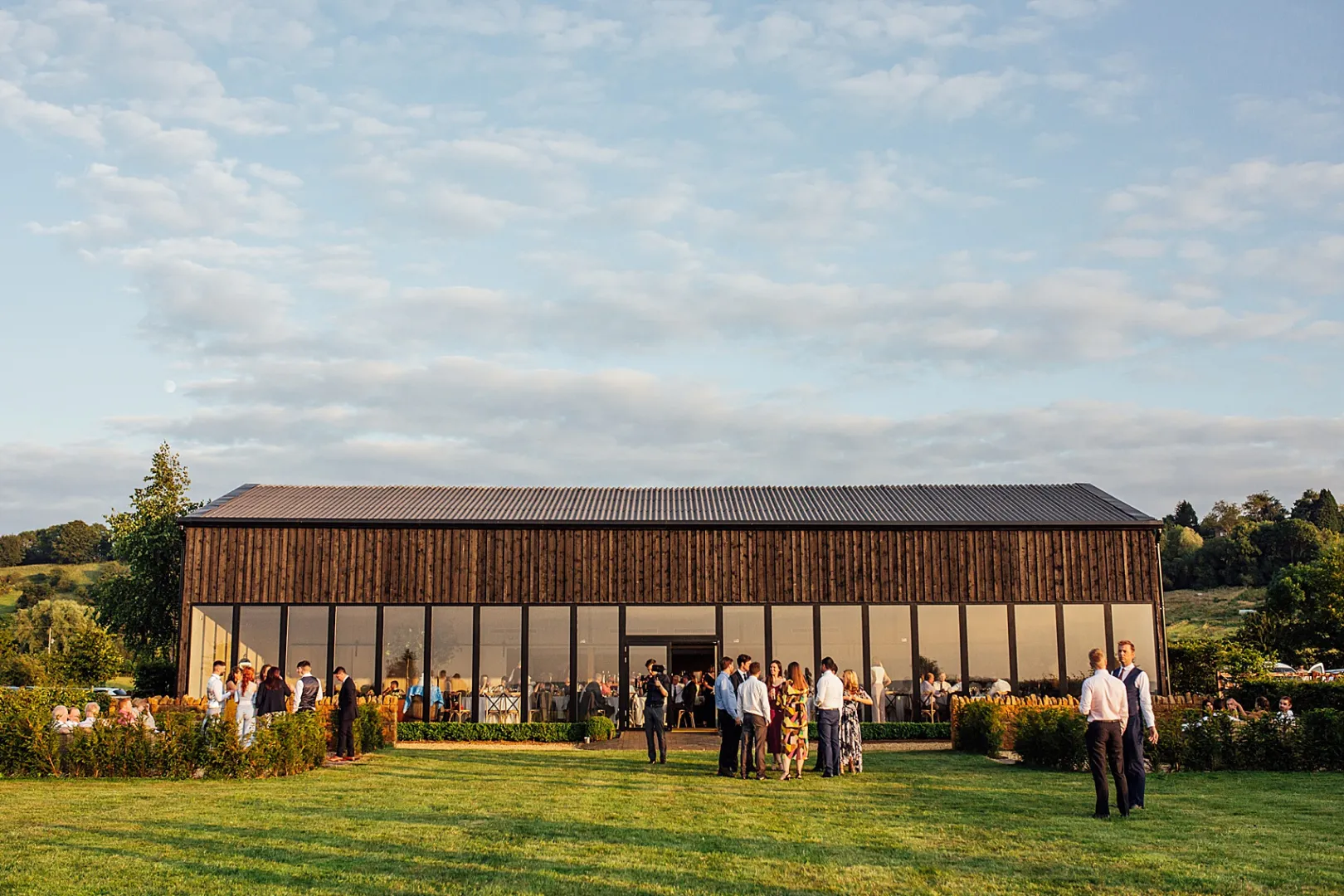 A modern rustic venue with floor-to-ceiling windows, surrounded by lush greenery and guests enjoying a scenic outdoor setting.