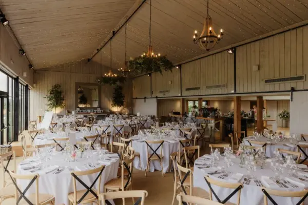 Spacious indoor reception area featuring elegant round tables, wooden accents, and large windows allowing natural light. Ideal for wedding celebrations.