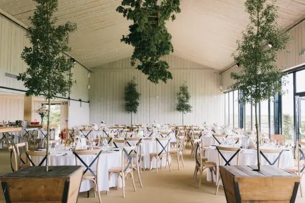 Bright, airy venue featuring elegantly set round tables, wooden accents, and lush greenery for a modern wedding atmosphere.