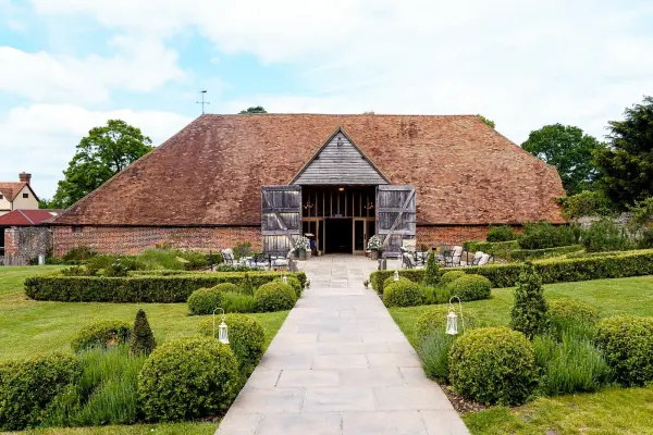 Charming rustic barn venue with a thatched roof, surrounded by manicured gardens and a stylish stone walkway leading to the entrance.