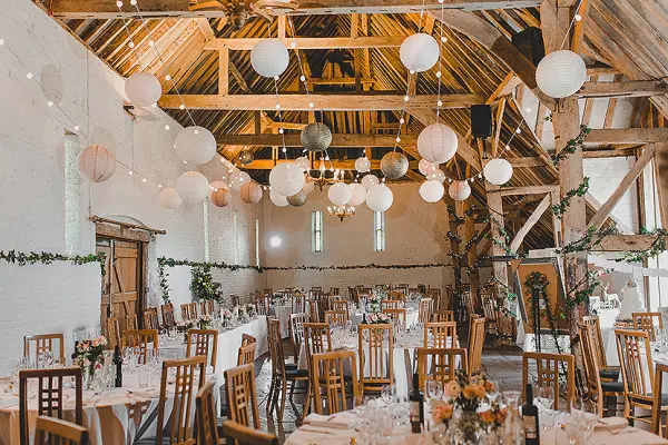Rustic barn venue featuring wooden beams, round tables, floral centerpieces, and elegant lanterns hanging from the ceiling.