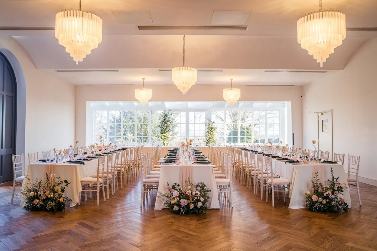 Bright ballroom with elegant chandeliers, long tables adorned with flowers, and large windows offering natural light and garden views.