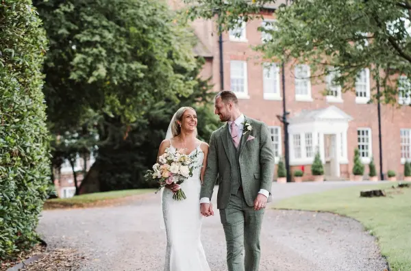 Bride and groom stroll hand in hand along a picturesque pathway, surrounded by lush greenery and an elegant brick venue in the background.