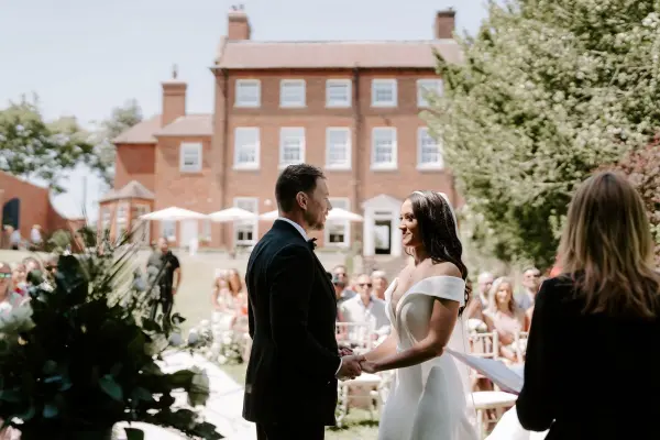 A charming outdoor ceremony features a couple exchanging vows, surrounded by lush gardens and a backdrop of a historic red brick manor.