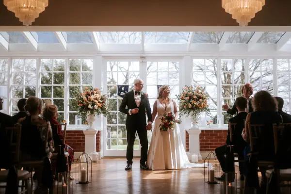 Bright and airy ceremony space with large windows, floral arrangements, and an elegant wooden floor. Couples share vows surrounded by guests.