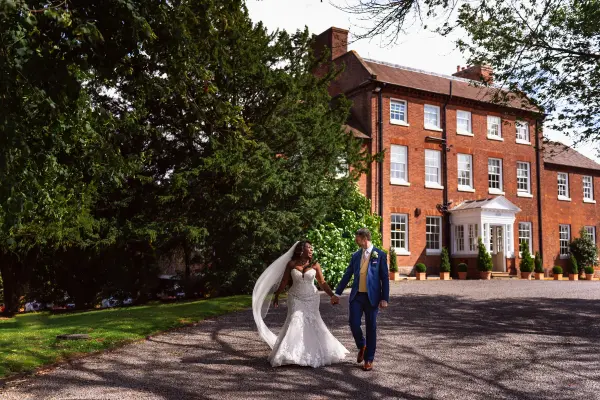 A couple strolls hand in hand along a scenic pathway, framed by lush greenery and a grand red brick manor house.