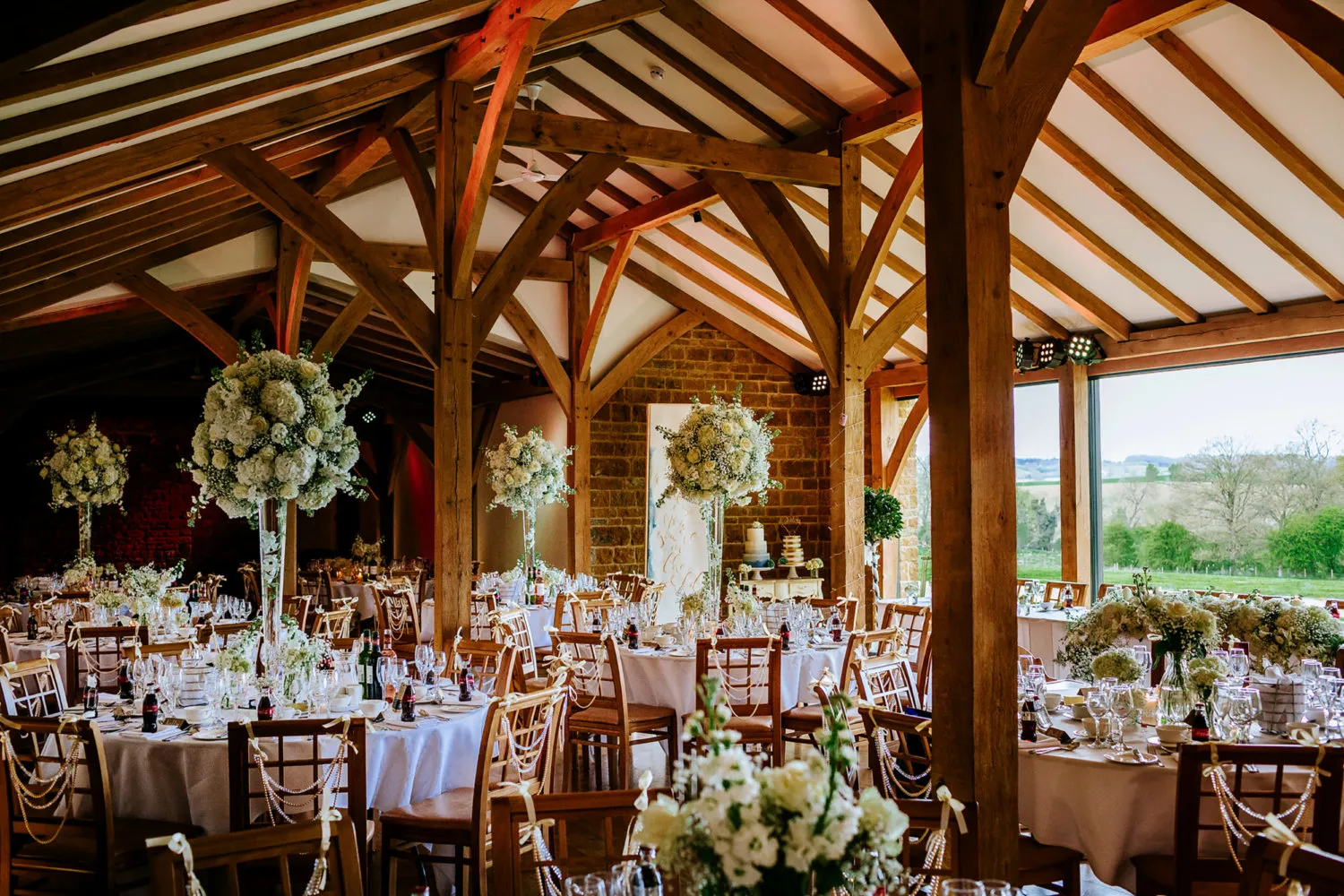 Elegant rustic venue with wood beams, round tables adorned with floral centerpieces, and panoramic views through large windows.