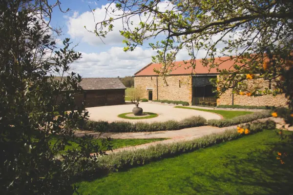 Charming rustic barn wedding venue with landscaped grounds, a circular driveway, and a central sculpture surrounded by greenery.