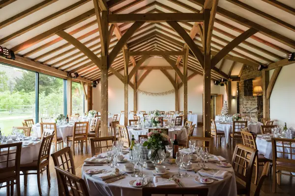Rustic barn interior featuring elegant round tables adorned with floral centerpieces, wooden beams, and large windows overlooking lush greenery.