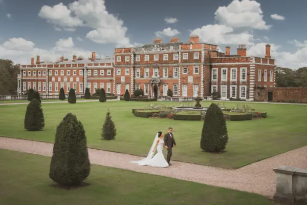 A couple strolls through manicured gardens, featuring elegant hedges and a grand mansion backdrop, perfect for a romantic wedding setting.