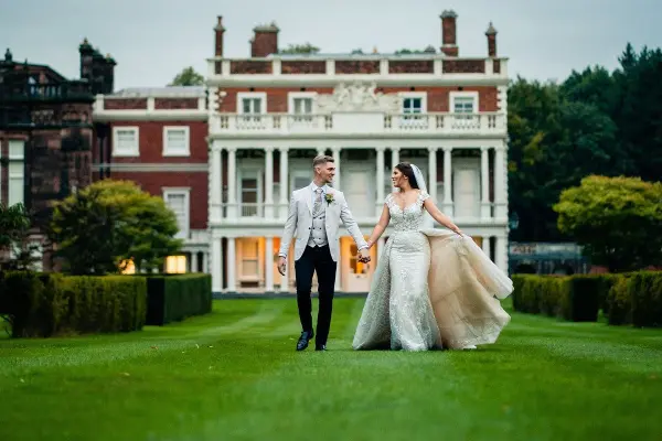 Couple walks hand in hand across a lush lawn, framed by a grand historic mansion and landscaped gardens, perfect for elegant weddings.