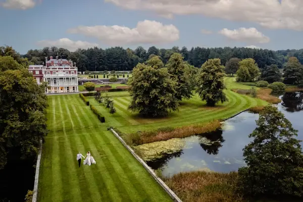 Bride and groom stroll through lush, manicured gardens beside tranquil water, framed by towering trees and a classic estate.