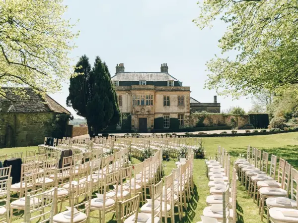 Charming outdoor ceremony space with elegant chiavari chairs set on lush grass, overlooking a grand manor house surrounded by greenery.