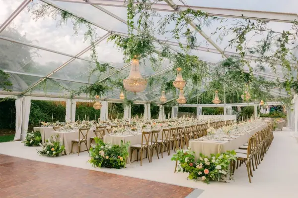 Elegant clear tent reception featuring long tables adorned with floral centerpieces and hanging lanterns, surrounded by lush greenery.
