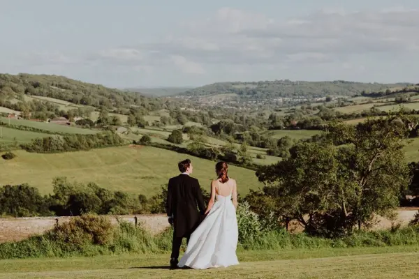 Couple stands hand-in-hand on a lush hillside, overlooking rolling green fields and a picturesque valley, perfect for a romantic outdoor wedding.