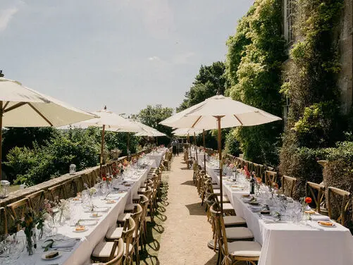 Elegant outdoor dining area featuring long tables adorned with flowers, set beneath stylish umbrellas amidst lush greenery. Perfect for alfresco celebrations.
