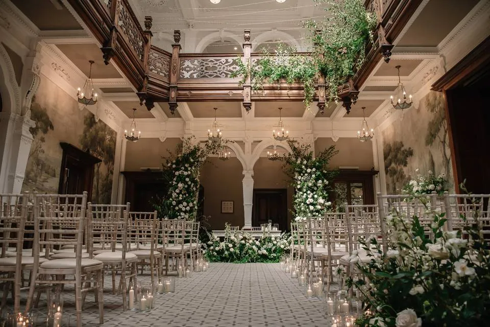 An elegant indoor wedding ceremony space featuring ornate wooden details, lush greenery, floral arrangements, and softly lit candles.