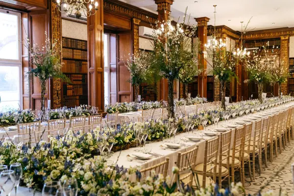 Elegant dining space featuring long tables adorned with lush floral centerpieces, surrounded by wooden bookshelves and chandeliers.