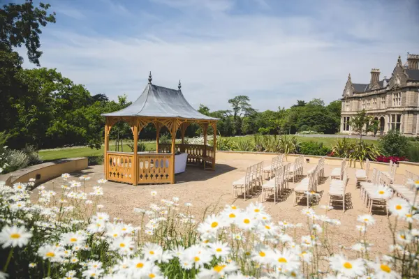 Charming outdoor ceremony space featuring a wooden gazebo surrounded by daisies and paved seating for guests, with a stately mansion in the background.
