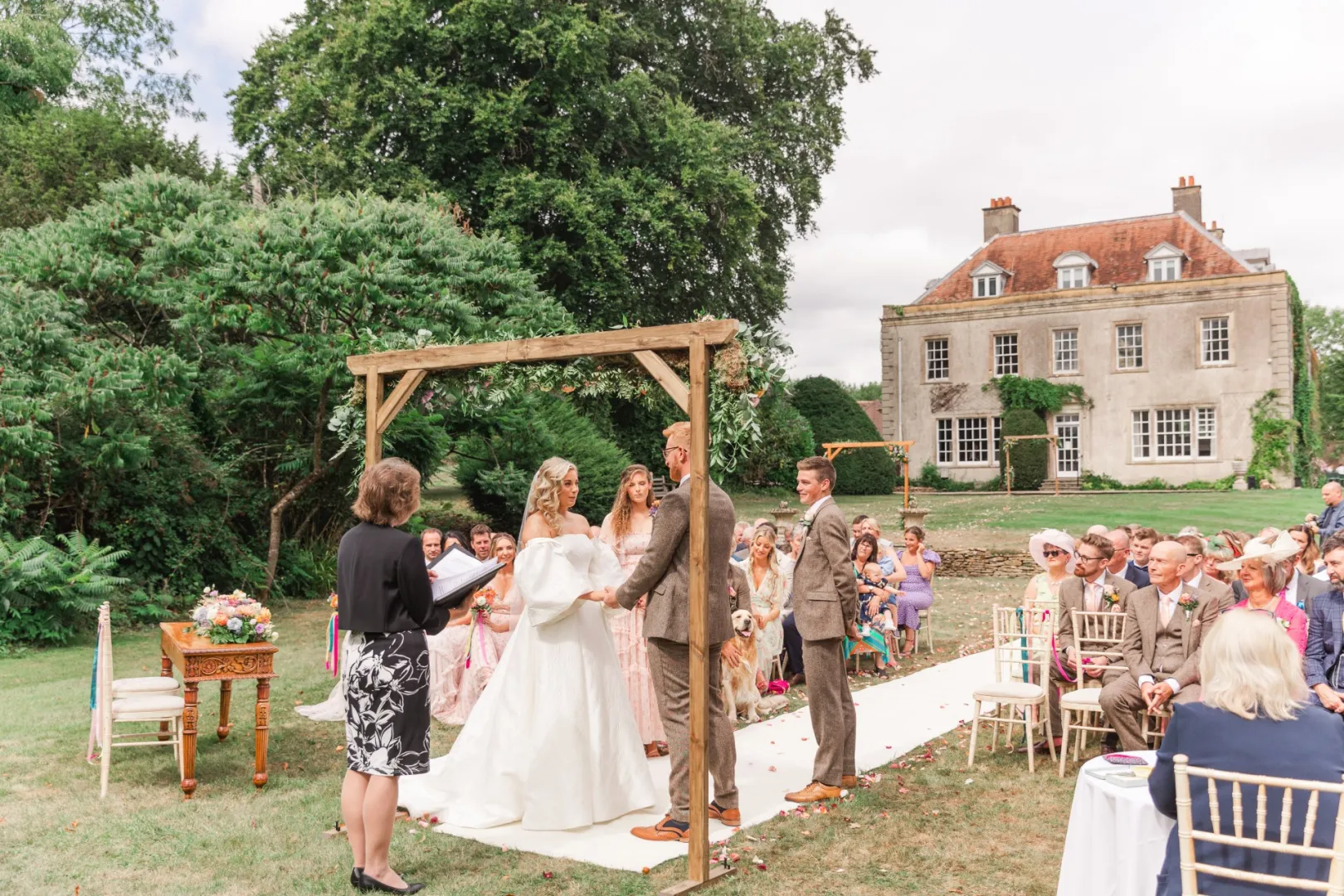 A charming outdoor ceremony under an elegant wooden arch, surrounded by lush greenery and a historic estate in the background.