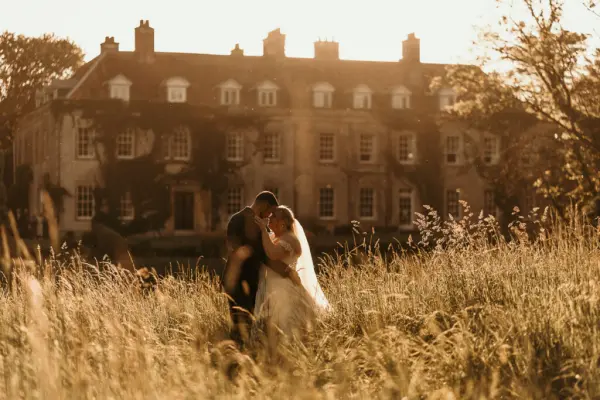 Couple embraces in a sunlit field near a historic estate, featuring lush greenery and elegant architecture, perfect for a romantic wedding.