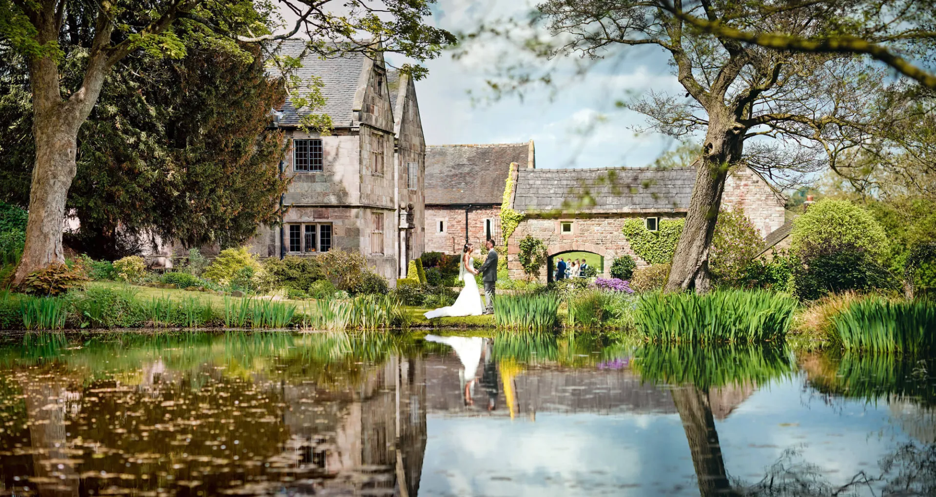 Charming wedding venue featuring a serene pond, lush greenery, and historic stone buildings, perfect for outdoor ceremonies.