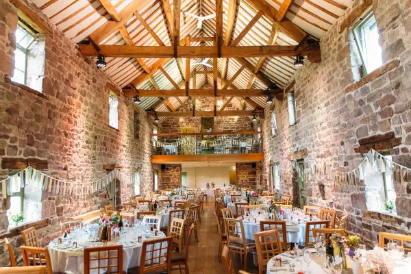 Charming rustic venue with exposed stone walls, wooden beams, and elegantly set tables for a romantic wedding reception. Bright windows add warmth.