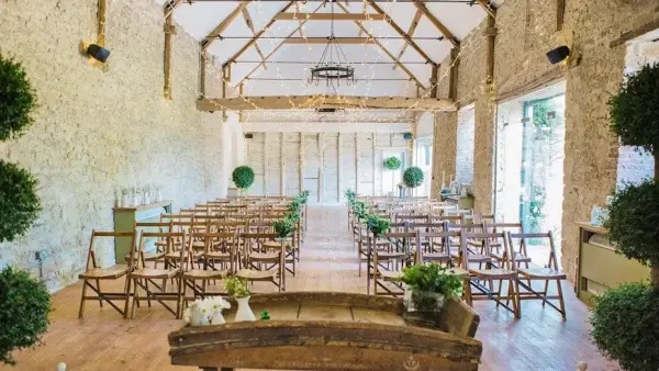 Rustic ceremony space featuring wooden beams, natural stone walls, and elegant greenery, with arranged chairs and warm wooden flooring.