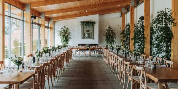 Bright and airy reception space featuring rustic wooden beams, long tables adorned with greenery, and large windows allowing natural light.