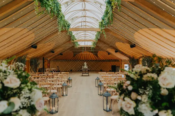 A beautifully designed indoor ceremony space features wooden beams, lush greenery, and rows of elegant chairs, creating a charming ambiance.