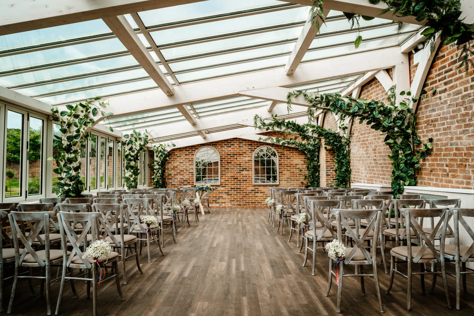 Bright glass-enclosed ceremony space featuring rustic brick walls, wooden floors, and floral-decorated arches for a romantic atmosphere.