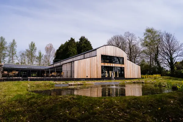 Modern barn venue with large glass windows, reflecting on a serene pond, surrounded by lush greenery and mature trees. Perfect for unique weddings.