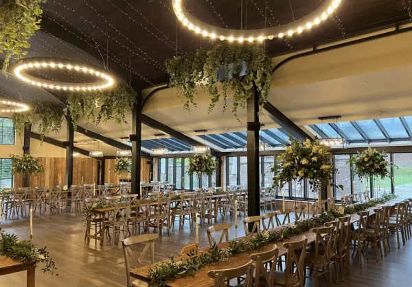Spacious venue featuring wooden tables, rustic chairs, and elegant greenery, with dramatic ring lighting and large windows for natural light.