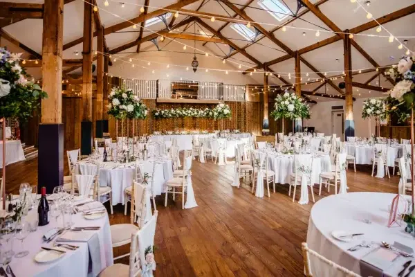 Rustic reception hall with wooden beams, elegant round tables, lush floral arrangements, and string lights creating a warm ambiance.